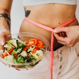 Woman holding vegetable salad and measuring waist with tape, symbolizing healthy lifestyle choices.
